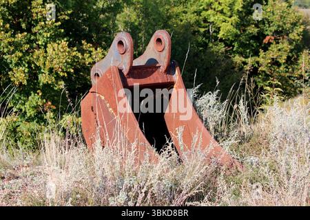 Grand godet de pelle rouillé se tient debout parmi les hautes herbes sèches et les plantes sauvages, abandonnés au bord d'un champ avec des arbres verts denses dans la BA Banque D'Images
