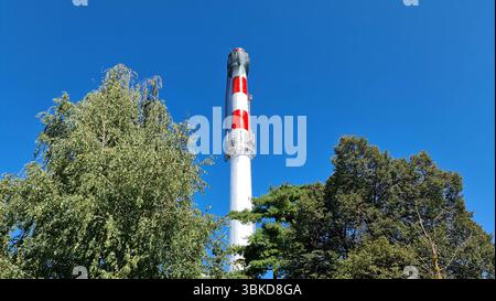 Une grande cheminée industrielle peinte en rayures rouges et blanches s'élève au-dessus des arbres feuillus, avec un échafaudage et un filet près du sommet, sur un bleu éclatant Banque D'Images