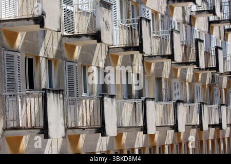 Des rangées de petits balcons avec des garde-corps en métal et des volets fanés créent un motif géométrique sur une façade ancienne d'appartement, projetant des ombres nettes en fort Banque D'Images
