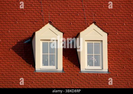 Deux lucarnes blanches aux fronts triangulaires se détachent sur un toit en tuiles rouges abruptes, projetant des ombres nettes, avec des vitres reflétant la lumière du soleil Banque D'Images