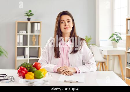 Portrait de jeune jeune amicale joyeuse femme médecin nutritionniste assis sur le lieu de travail dans le bureau. Banque D'Images