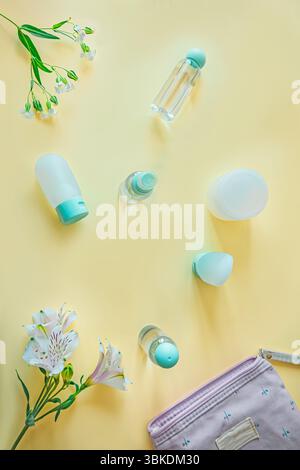 Pose plate verticale de bouteilles de toilette de voyage, pochette florale et délicates fleurs blanches sur fond pastel. Composition propre et féminine, idéale pour Banque D'Images
