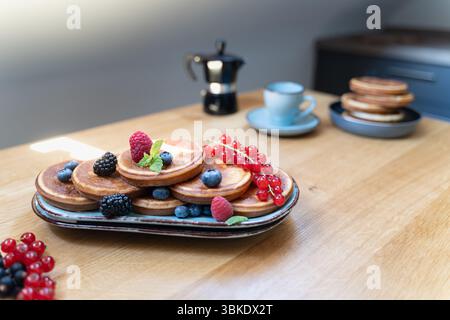 Crêpes surmontées de baies et de tranches de banane sur une assiette bleue, café et pile supplémentaire en arrière-plan, lumière naturelle du dessus. Banque D'Images