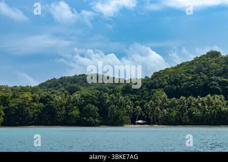 Une île luxuriante et verdoyante s'élève des eaux turquoises, sa végétation dense contraste vibrant avec le ciel clair au-dessus. Une petite structure nichée parmi les palmiers sur le rivage, offrant un aperçu de la présence humaine au milieu de la beauté naturelle. Banque D'Images