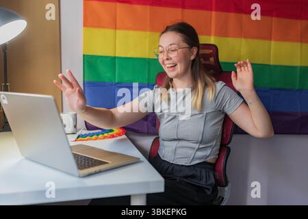 Heureuse femme saluant pendant la réunion vidéo, drapeau arc-en-ciel toile de fond visible / souriante jeune femme levant la main devant l'ordinateur portable, assise au bureau avec RA Banque D'Images