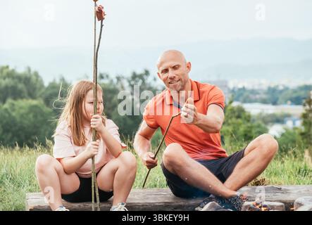 Scène extérieure de famille mignonne du père montrant sa fille comment mettre la saucisse sur le bâton de barbecue alors qu'ils sont assis à côté d'un feu de camp pendant la randonnée dans la nature e Banque D'Images
