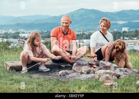 Scène extérieure mignonne de pique-nique : père, fille et maman avec chien Maltipoo griller des saucisses sur des bâtons de barbecue assis près du feu de camp pendant la randonnée R Banque D'Images