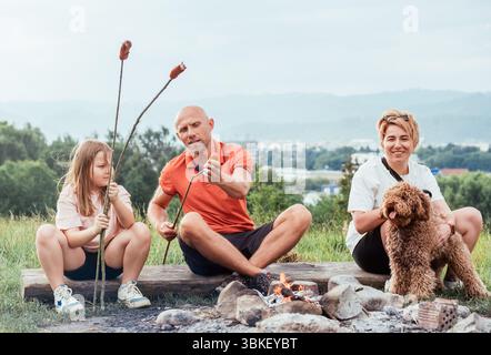 Père montrant à sa fille comment mettre la saucisse sur le bâton de barbecue tout en étant assis près du feu de camp avec la mère et le chien Maltipoo pendant la randonnée reflétant fami Banque D'Images