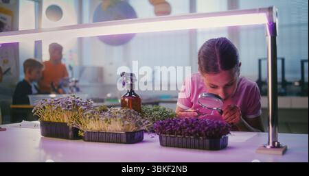 Fille talentueuse assise à table et utilisant un smartphone, menant une expérience de biologie sur les plantes avec une pince à épiler et une loupe. Enfants de l'école primaire pendant la leçon en classe. Éducation STEM. Banque D'Images