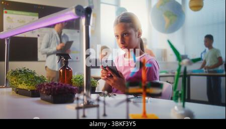 Fille talentueuse assise à table et utilisant un smartphone, menant une expérience de biologie sur les plantes avec une pince à épiler et une loupe. Enfants de l'école primaire pendant la leçon en classe. Éducation STEM. Banque D'Images