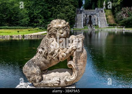 Un lion de pierre sur une jetée de pont dans les jardins classés du Château de la Roche Courbon. En arrière-plan, l’étang du jardin et l’escalier d’eau en pierre avec son architecture à baldaquin, qui marque la fin de l’axe du jardin. Rue de la belle au bois dormant, Saint-Porchaire, Charente-maritime, Nouvelle-Aquitaine, France Banque D'Images