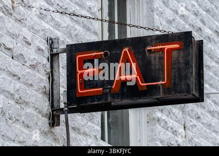 Une enseigne rouge vif au néon affichant le mot « EAT » est montée sur un vieux bâtiment en briques. Le cadre en métal vieilli et le mur en pierre texturée ajoutent un aspect rugueux et urbain Banque D'Images