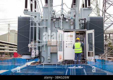 Un ingénieur effectue une inspection sur un grand transformateur à la sous-station extérieure assurant la sécurité et la fonctionnalité dans un environnement industriel avec protection Banque D'Images