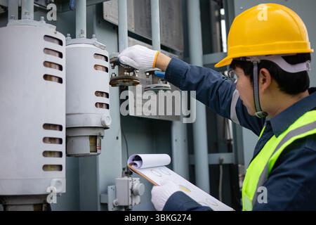 Un ingénieur portant un casque jaune effectue l'inspection du transformateur à la sous-station tout en tenant un presse-papiers avec des graphiques et des données pour des évaluateurs précis Banque D'Images