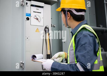 Un ingénieur portant un casque jaune effectue l'inspection du transformateur à l'intérieur de la sous-station avec une attention ciblée et un équipement de sécurité pour une évaluation précise Banque D'Images