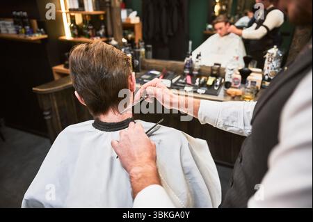 Gros plan d'un coiffeur expérimenté à l'aide d'un peigne et de ciseaux pour couper les cheveux supplémentaires. Jeune client pour la première fois dans un salon de coiffure. Vue arrière du travail du coiffeur. Banque D'Images