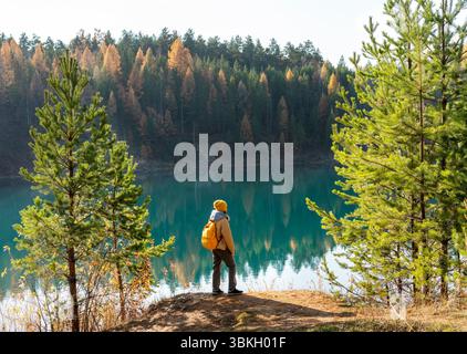Jeune femme touristique avec sac à dos jaune marchant près du lac contre la forêt de pins d'automne regardant la randonnée de vue Banque D'Images