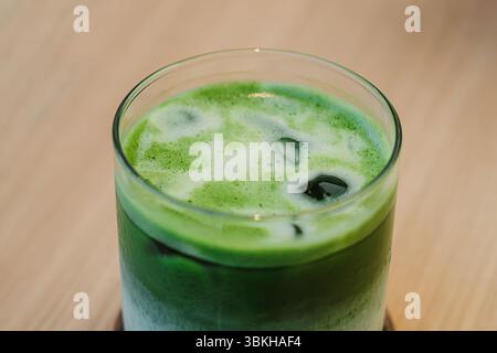 Un verre de latte matcha glacé sur un dessous de verre en bois se trouve sur une table en bois clair Banque D'Images
