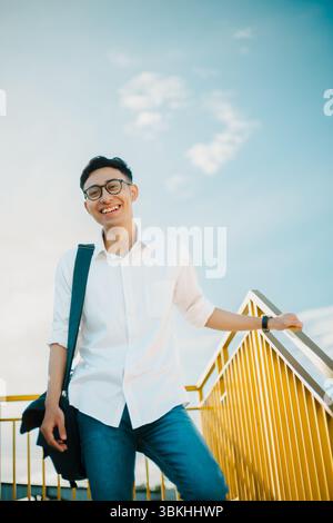 L'homme d'Asie du Sud-est sourit tout en se tenant debout sur un escalier jaune contre un ciel bleu brillant sous le pont suspendu Kampung Warna-Warni Jodipan, Malang Banque D'Images