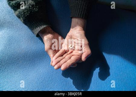 Top shot de mains seniors tenant des pilules de médicaments sur un tapis bleu, éclairé par la lumière naturelle du soleil Banque D'Images