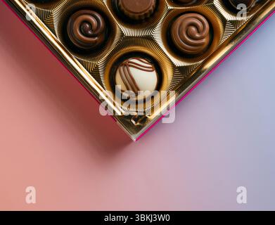 Gros plan du coin d'une boîte en or remplie de divers chocolats, présentant des motifs complexes et une bordure rose sur fond violet Banque D'Images
