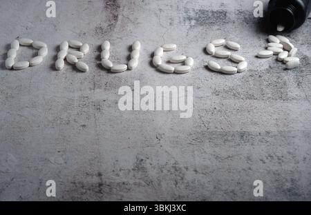 Les pilules ovales blanches se déversent d'une bouteille renversée à côté du mot « drogues » formé par des comprimés sur une surface grise texturée. Overdose et addiction Banque D'Images
