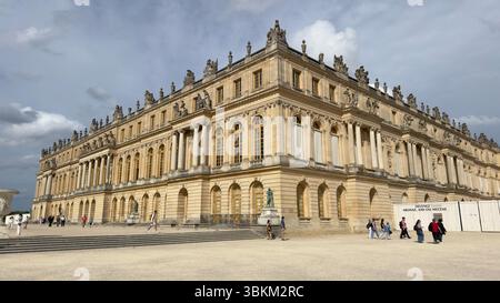 Vue extérieure du château de Versailles avec les gens, les escaliers et un ciel nuageux. Bâtiment jaune pâle avec de nombreuses fenêtres et colonnes. Banque D'Images