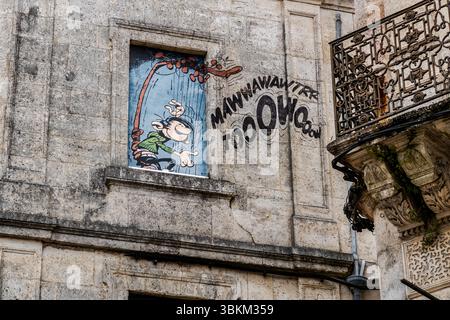Dans la fenêtre supérieure d'une fresque, le messager de bureau Gaston joue du gaffophone, une sorte de harpe qui produit des bruits absolument désagréables, Rue Hergé, Angoulême, Nouvelle-Aquitaine, France. Gaston et Prunelle sont des personnages de la bande dessinée belge créée par André Franquin. Le nom original de Gaston est "Gaston Lagaffe", et il est un messager de bureau chaotique qui provoque de nombreux accidents. Le son peint "MAWWAWAWTRR TOOOWOOOW" s'étend sur la façade en pierre. La fresque date de 1999. À droite, un balcon en fer forgé dépasse sur des corbeilles en pierre ornées. La ville est une Ville créative de l'UNESCO de Li Banque D'Images