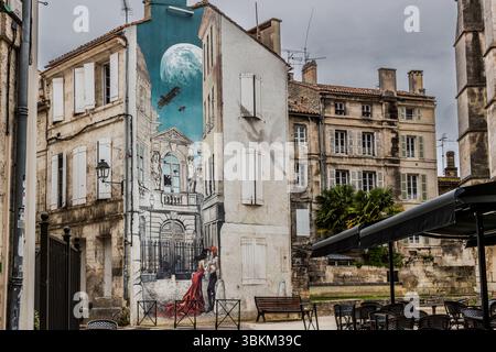 Une murale représente une scène avec un couple, un avion et une lune sur fond d'architecture historique. C’est un exemple de Street art qui embellit la ville et reflète l’histoire et la culture de la région. Angoulême est reconnue comme ville créative de l’UNESCO dans la catégorie littérature depuis 2019. Rue Sainte-Marie, Angoulême, Nouvelle-Aquitaine, France Banque D'Images