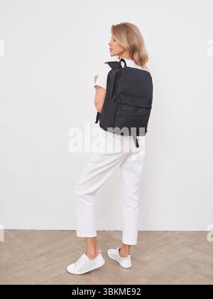 Jeune femme aux longs cheveux blonds portant un t-shirt blanc décontracté et un pantalon, debout sur le côté avec un sac à dos noir, regardant à gauche. Elle est en blanc Banque D'Images