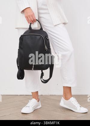 Femme à la mode tient élégant sac à dos noir contre un pantalon blanc et des baskets, mettant en valeur une tenue décontractée élégante. Le parquet contraste avec elle Banque D'Images