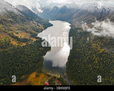 Une vue aérienne montre les eaux réfléchissantes du lac Rakhmanovskoe niché dans une vallée entourée de forêts d'automne et de montagnes enneigées Banque D'Images