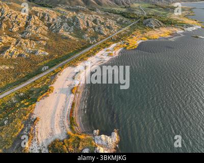 Une vue aérienne met en valeur un rivage accidenté du lac avec une étendue de plage de sable, bordée par un terrain rocheux et des montagnes lointaines sous une lumière douce Banque D'Images