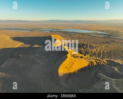 Une vue aérienne capture la forme dramatique de la dune de sable Singing Barkhan projetant une longue ombre à travers le paysage désertique au coucher du soleil, avec w lointain Banque D'Images