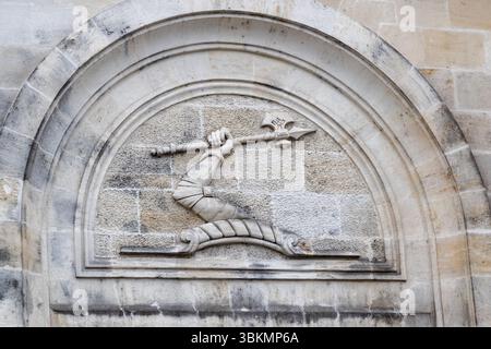 Le symbole « bras armé » au-dessus du portail principal de la maison de cognac Hennessy à Cognac provient des armoiries du fondateur Richard Hennessy et représente un bras blindé tenant une halberde (souvent représentée comme une hache). Façade du bâtiment historique Hennessy & Co. avec son entrée distinctive et l'inscription 'Jas Hennessy & Co.'. Il témoigne de la riche histoire et du patrimoine de la production de cognac dans la région. Le détail d'un relief montre un bras tenant une halberde, encastré dans une sculpture décorative en pierre voûtée. Quai Maurice Hennessy, Cognac, Nouvelle-Aquitaine, France Banque D'Images