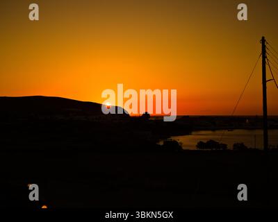 Coucher de soleil orange ardent sur Apollonia Bay sur l'île de Milos, avec des silhouettes de collines côtières. Banque D'Images