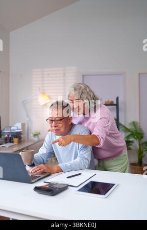Un couple asiatique plus âgé partage des moments joyeux à la maison tout en utilisant un ordinateur portable ensemble. Ils apprécient les activités en ligne et montrent de l'amour tout en tissant des liens Banque D'Images