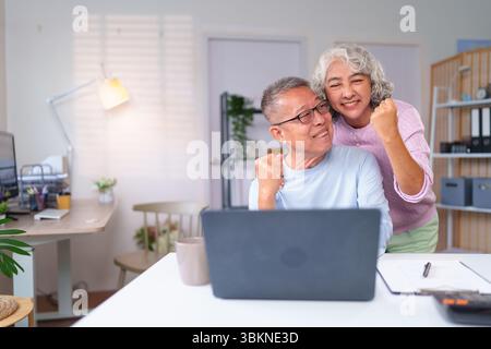 Un couple asiatique plus âgé célèbre son succès avec un ordinateur portable à la maison. Ils profitent de leur heureuse vie de retraite ensemble. Ils sont contents Banque D'Images