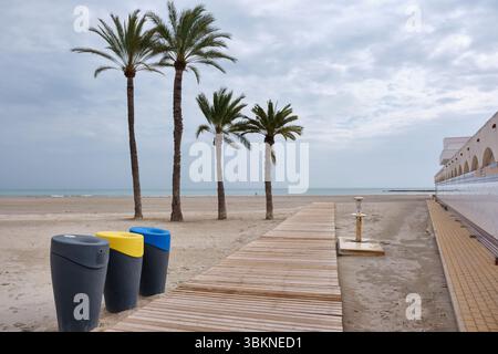Scène de plage calme à Santa Pola, province d'Alicante avec des palmiers, une promenade en bois et des poubelles colorées sous un ciel nuageux le long de la côte Banque D'Images
