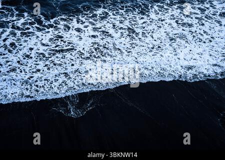 Une vague mousseuse s'écrase sur une plage de sable sombre, créant une scène sereine et naturelle. Le contraste entre la mousse blanche et le sable foncé crée une visualisation Banque D'Images