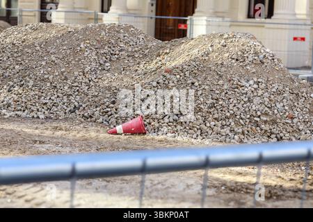 Scène d'un chantier de construction avec un gros tas de gravier ou de gravats sur la droite, un cône rouge de circulation au premier plan, et une route avec véhicule Banque D'Images