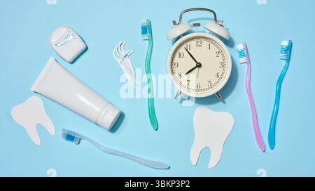 Tube blanc de dentifrice, fil dentaire dans un étui, brosses à dents de différentes couleurs, réveil blanc, soie dentaire en plastique placée sur un fond bleu. T Banque D'Images
