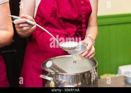 Gros plan d'une femme dans un tablier rouge à l'aide d'une louche pour verser la pâte crémeuse dans une tasse en aluminium à partir d'une grande casserole. Banque D'Images