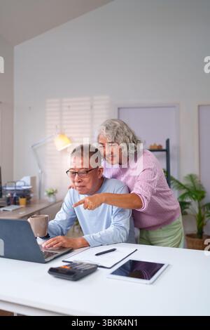 Un couple asiatique plus âgé partage des moments joyeux à la maison tout en utilisant un ordinateur portable ensemble. Ils apprécient les activités en ligne et montrent de l'amour tout en tissant des liens Banque D'Images