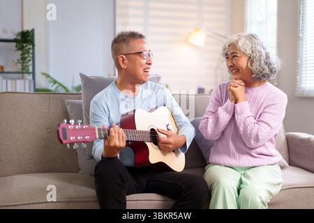 Un homme asiatique plus âgé joue de la guitare pour sa femme à la maison, ils sont assis sur le canapé et profiter de leur temps ensemble couple senior ayant du plaisir à l'intérieur Banque D'Images