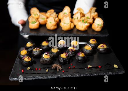 Assortiment de canapés élégants servis sur des plateaux en ardoise noire, y compris des feuilletés salés noirs et des pâtisseries fourrées à choux, idéales pour les événements. Banque D'Images