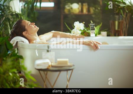 Une femme adulte aux cheveux foncés se détend dans un bain blanc moussant, les yeux fermés avec un sourire serein. Entouré de plantes vertes luxuriantes et d'un éclairage doux, embodi Banque D'Images