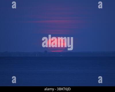 Strawberry Moon se lève avec un voile léger, rouge et grand, sur le Greifswalder Bodden le soir. Copier l'espace. Espace pour textes et desi Banque D'Images