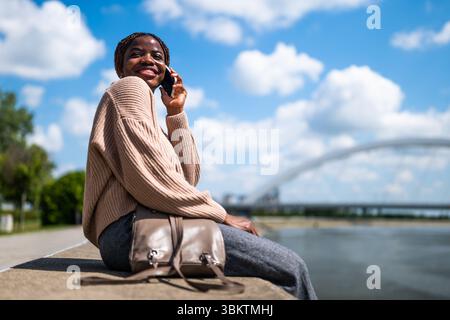 Une jeune femme noire joyeuse parle au téléphone. Elle rit et profite d'une journée ensoleillée dans la ville. Banque D'Images