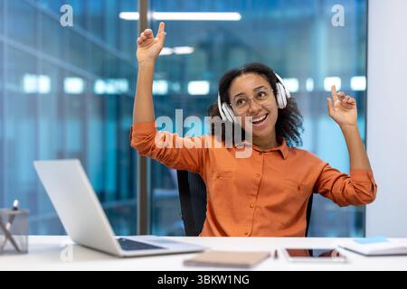 Une jeune femme joyeuse portant des écouteurs et des lunettes, célébrant dans un cadre de bureau avec un ordinateur portable. Banque D'Images